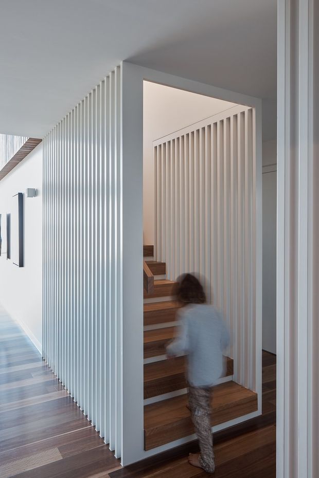 Interior architectural photograph of a bright, minimalist stairway enclosed by white vertical slatted screens and framed openings. Warm timber stair treads and dark timber flooring contrast with the white walls and balustrade-like partitions. A blurred child is moving on the stairs, adding a sense of residential use and depth.