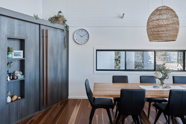 A bright contemporary dining room with a large timber dining table and black upholstered dining chairs. The room features dark blue-grey built-in cabinetry with open shelving and tall vertical timber handles, white horizontal wall panelling, warm timber flooring, a narrow horizontal window, and a large woven pendant light. Minimal decorative styling and indoor plants add natural texture.