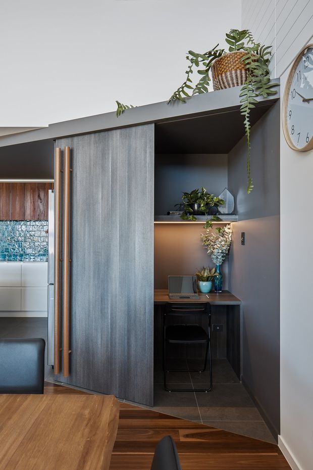 A compact built-in home office nook beside a dining area, partially screened by a tall dark timber door with long vertical timber handles. The workspace includes a fixed dark desk, black chair, laptop, shelving with integrated warm lighting, and decorative plants. The surrounding interior features dark grey cabinetry or panelling, a warm timber dining table and floor, white vertical wall panelling, and a partially visible kitchen beyond.