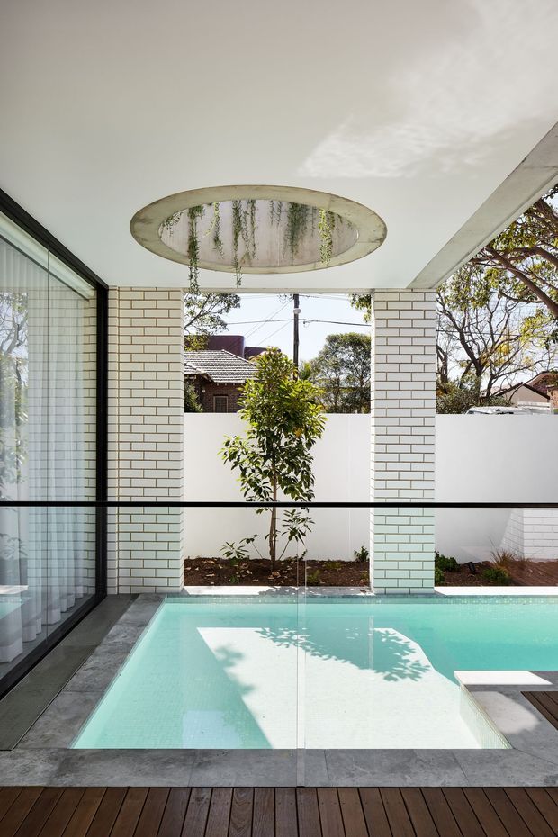 A bright, modern indoor-outdoor pool courtyard viewed from beneath a covered terrace. The scene features a turquoise plunge pool, dark timber decking, grey stone or concrete pool surrounds, white glazed brick columns and walls, full-height black-framed glazing, and a distinctive circular ceiling opening with hanging greenery. A small planted courtyard and neighbouring residential context are visible beyond.