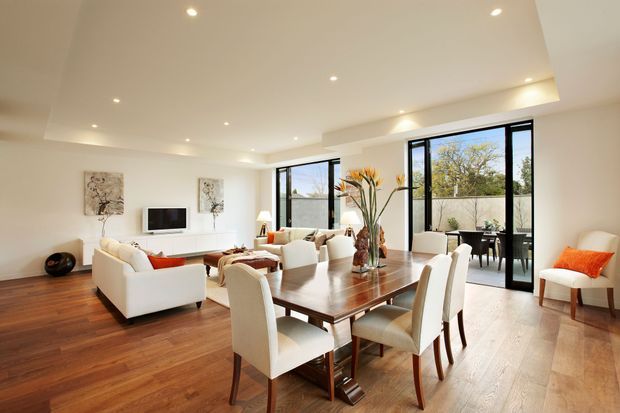 Bright open-plan living and dining area with warm timber flooring, a large polished wood dining table surrounded by upholstered chairs, and a white seating arrangement facing a wall-mounted television. Floor-to-ceiling black-framed glazing opens toward a small outdoor terrace, while recessed ceiling lights and neutral walls create an airy, refined interior.