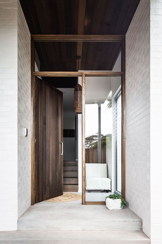 A sheltered residential entry porch with white-painted brick walls, a tall glazed timber-framed sidelight and transom, and a dark timber pivot door. The recessed entry has a dark timber-lined ceiling, concrete steps and floor, a woven pendant light, and a glimpse of an interior stairway and seating beyond the doorway. The composition is warm, refined, and strongly material-focused.