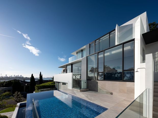 A contemporary multi-level white residence with extensive floor-to-ceiling glazing, dark window frames, angular roof and facade geometry, and a pale stone-clad terrace. The view looks across a landscaped sloping site toward a distant urban skyline, with a reflecting swimming pool in the foreground and glass balustrades along the terrace and stairs.