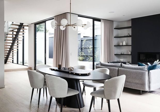 Bright open-plan contemporary interior featuring a dining area with a black oval table and upholstered chairs, opening into a living room with a large grey sectional sofa. Full-height black-framed glazing, sheer curtains, built-in open shelving, a sculptural pendant light, and an open staircase are visible.