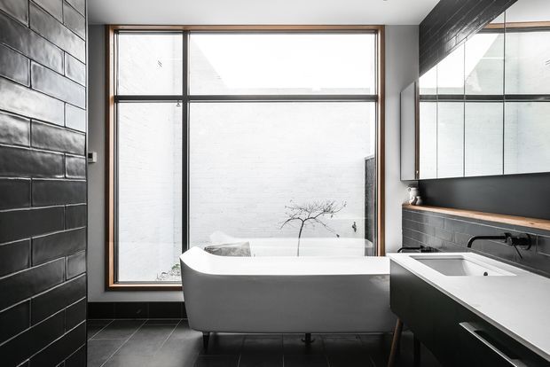 A contemporary bathroom featuring a freestanding white bathtub positioned beside a large black-framed window, a dark tiled feature wall, matching dark floor tiles, and a long floating vanity with a white integrated basin. A wall-to-wall mirror, black tapware, and timber-toned vanity shelf add contrast. The room has a clean, monochromatic and refined aesthetic with strong natural light.