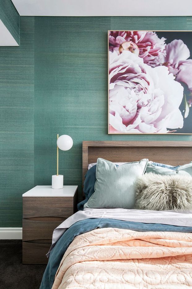 A stylish bedroom vignette featuring a timber bed with a simple integrated headboard, layered blue, blush and pale cushions and bedding, and a dark wood bedside table with a white top. The room has deep teal textured wallcovering, a slim brass-and-white globe table lamp, and a large framed floral artwork above the bed. The palette is rich yet soft, with a refined contemporary feel.