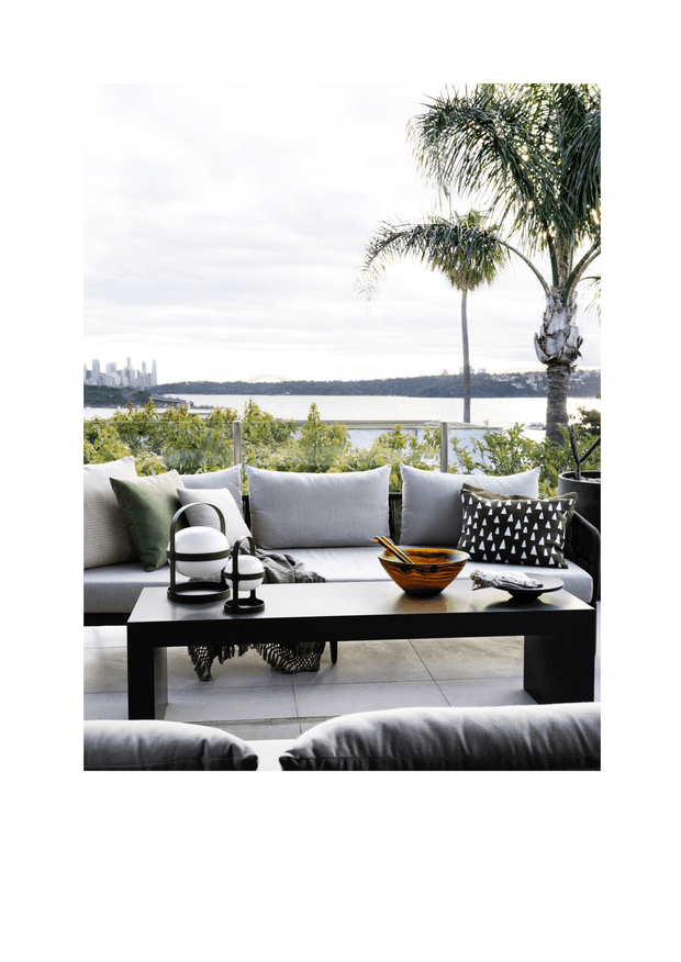 A bright, sophisticated outdoor terrace lounge overlooking a wide body of water and a distant urban skyline. The space features a large light-grey upholstered sofa with patterned and solid cushions, a black rectangular coffee table, decorative ceramic and sculptural objects, pale tiled flooring, glass balustrading, lush planting, and tall palms. The setting feels relaxed yet refined, with strong indoor-outdoor styling and expansive waterfront views.