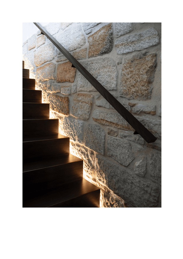 Close-up architectural photograph of a dark timber staircase ascending beside a rugged, irregular natural stone masonry wall. A slim dark handrail runs diagonally along the wall, while warm concealed lighting washes across the stone and stair edges, creating a dramatic textured effect.