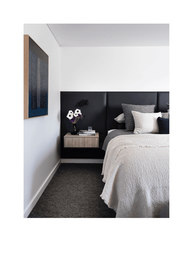 A refined modern bedroom with a partially visible bed, dark upholstered full-height headboard, floating timber bedside table, black wall-mounted reading light, textured neutral bedding, grey carpet, white walls, and abstract dark-toned artwork.