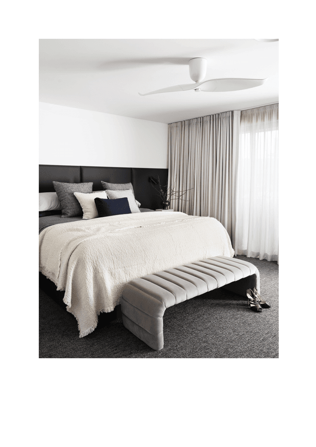 A bright, refined contemporary bedroom featuring a large bed with a black upholstered headboard, layered neutral bedding and cushions, a softly upholstered channel-tufted bench at the foot of the bed, full-height beige curtains with sheer drapes, and dark grey carpet. A white ceiling fan provides a streamlined overhead feature.