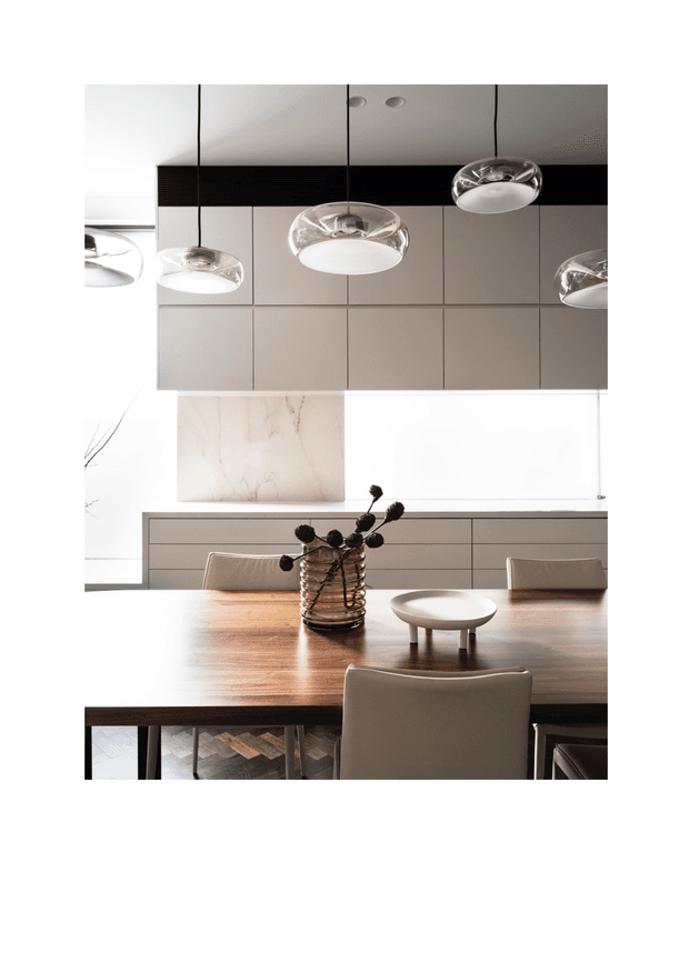 A refined contemporary dining area with a large dark timber dining table, upholstered dining chairs, a sculptural glass vase, and a small round centrepiece. The room features pale integrated cabinetry, a large light stone or marble panel, dark horizontal detailing, and multiple suspended glass pendant lights. The palette is warm and neutral with strong geometric lines and abundant natural light.