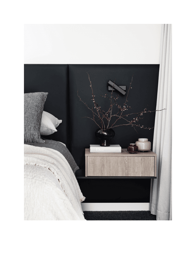A close-up view of a refined bedroom corner featuring a bed with a tall black upholstered headboard, layered grey and white bedding, a floating light timber bedside cabinet, and minimalist wall-mounted artwork. A black vase with delicate branches and small decorative vessels sit on the cabinet. White curtains frame the right side, while dark carpet and a white skirting board complete the setting.