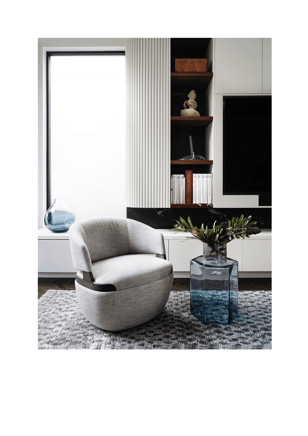 A refined contemporary living room vignette with a curved light-grey upholstered lounge chair positioned beside a transparent blue glass side table and vase. Behind it is a built-in media wall with white cabinetry, open dark timber shelving, a black marble-look feature band, television, books and sculptural decor. A tall window and vertically fluted wall panel add texture, while a grey patterned rug grounds the seating area.