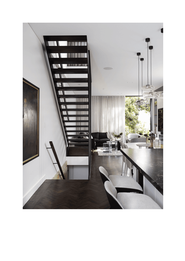 A refined open-plan interior combining a kitchen, dining and living area. A dramatic black open-riser staircase runs along a white wall, while a dark stone island with upholstered bar stools occupies the foreground. Dark herringbone timber flooring, sheer curtains, sculptural pendant lights and large glazing create a sophisticated, high-contrast space with views to greenery outside.