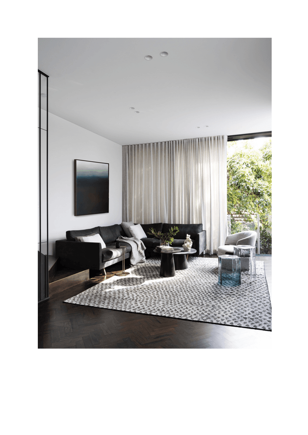 A refined contemporary living room with black upholstered sofas, a low black coffee table, sculptural decor, and a grey patterned area rug. Dark herringbone timber flooring contrasts with white walls and full-height sheer curtains. Large glazing brings in natural light and views of greenery, while recessed ceiling lights and a monochrome artwork complete the calm, sophisticated interior.