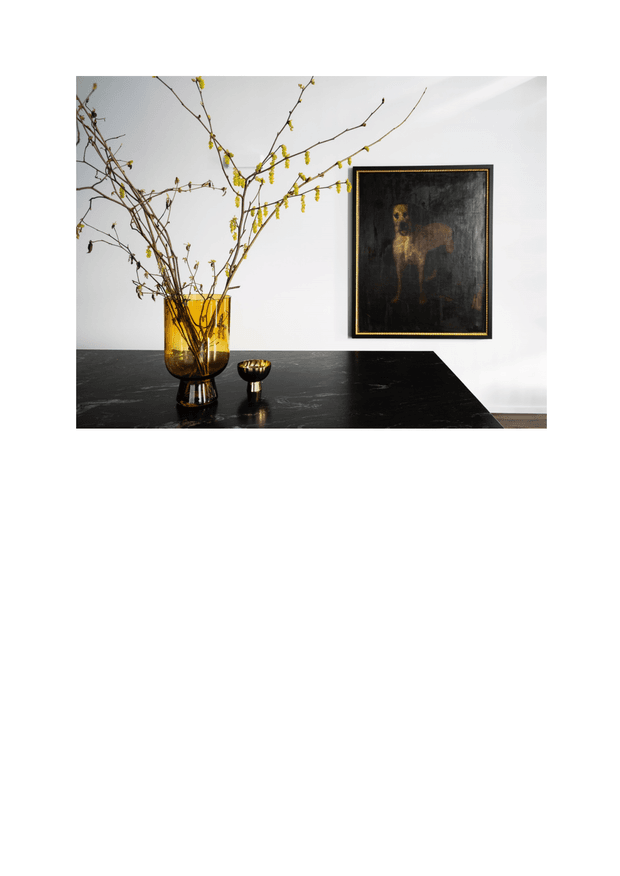 A refined interior vignette featuring a dark polished stone console or benchtop against a white wall. An amber glass vase with bare flowering branches and a small matching vessel sit on the surface, beside a dark framed artwork with a thin metallic border. The composition has a restrained, gallery-like atmosphere with strong black, white, and amber contrast.