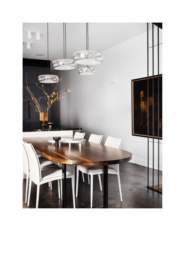 A refined contemporary dining area with a large oval dark timber dining table, upholstered white dining chairs, and sculptural glass pendant lights. The room features dark cabinetry and a dark stone kitchen counter in the background, pale walls, herringbone timber flooring, a black-framed glazed partition, and a dramatic framed artwork with warm metallic tones.