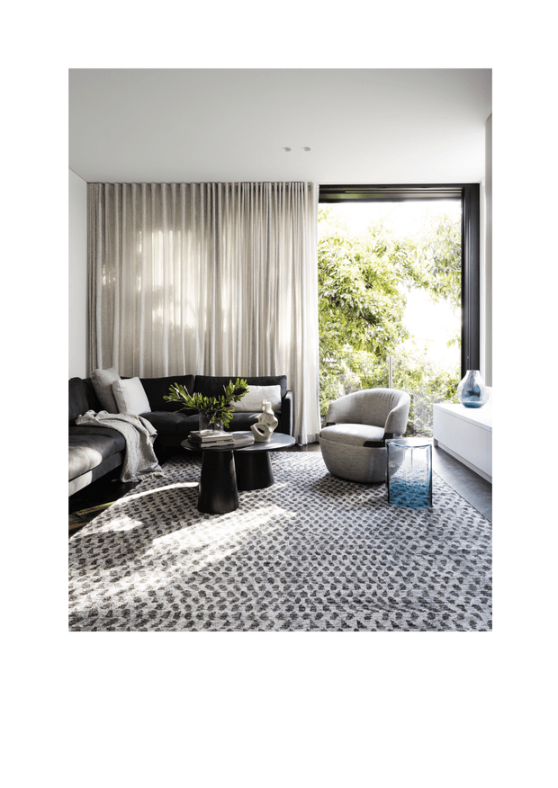 A bright, refined contemporary living room with a charcoal sectional sofa, round black coffee tables, a sculptural accent chair, a large textured grey rug, and a low white media console. Full-height sheer curtains cover one wall, while a large black-framed glazed opening brings in leafy outdoor views and strong natural light. The palette is predominantly neutral with subtle blue accents in the glass decor and side table.