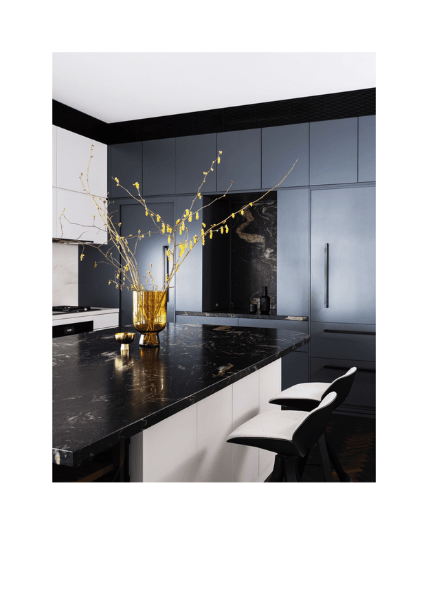 A refined contemporary kitchen featuring dark blue-grey full-height cabinetry, a dramatic black marble island benchtop with light veining, integrated appliances, and a dark marble backsplash. Two sculptural black-and-white counter stools sit at the island, while a gold-toned vase with branches provides a warm decorative accent. The space has a clean, luxurious and moody atmosphere.