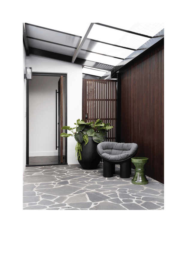 A refined covered entry courtyard with a translucent roof canopy, white rendered walls, dark timber slatted screening, and an irregular grey stone floor. The space includes a black planter with lush greenery, a sculptural charcoal lounge chair, and a small green side table, creating a contemporary, sheltered outdoor arrival area.