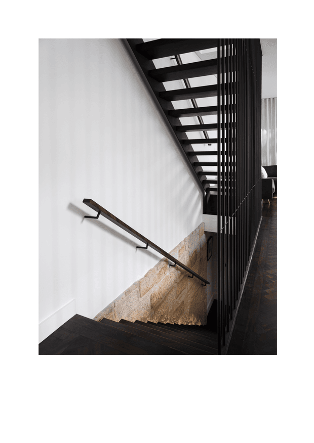 Interior architectural photograph looking down a modern stairway. The staircase has dark timber treads, a slim dark handrail, pale natural stone wall cladding along the lower stairwell, and a black vertical-slat balustrade or screen. A large skylight or glazed overhead opening brings daylight into the stairwell, with a glimpse of an adjacent dark-floored room beyond.