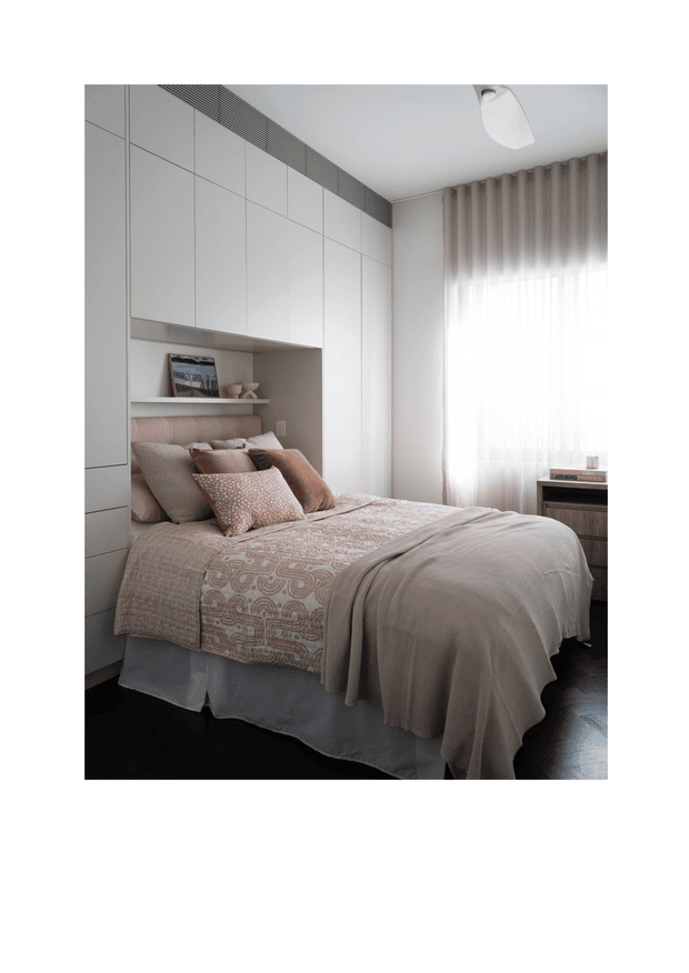 A compact, softly styled bedroom with a double bed dressed in layered beige and taupe bedding, patterned cushions, and a textured throw. Full-height white built-in cabinetry incorporates an open display niche behind the bed. Sheer curtains and taupe drapes filter daylight from the window, while a small dark bedside table and simple ceiling light complete the calm, contemporary space.
