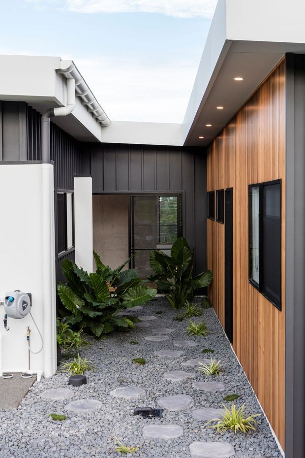 A narrow landscaped courtyard or side passage between modern residential wings. The space features grey gravel, irregular concrete stepping stones, low ornamental grasses and broad-leaf planting. Surrounding architecture combines black vertical cladding, warm timber-look vertical wall cladding, white rendered walls, black-framed windows, exposed guttering and a projecting flat-roofed eave with recessed downlights.