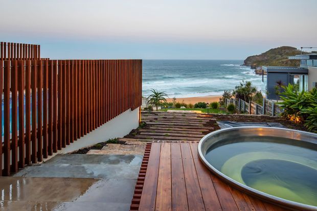 A contemporary coastal outdoor terrace overlooking the ocean, featuring a timber deck and stepped timber walkway, a curved plunge pool or spa, vertical timber privacy screening, concrete paving, and landscaped planting. A modern residence is partially visible on the right, with a sandy beach and rocky headland beyond.