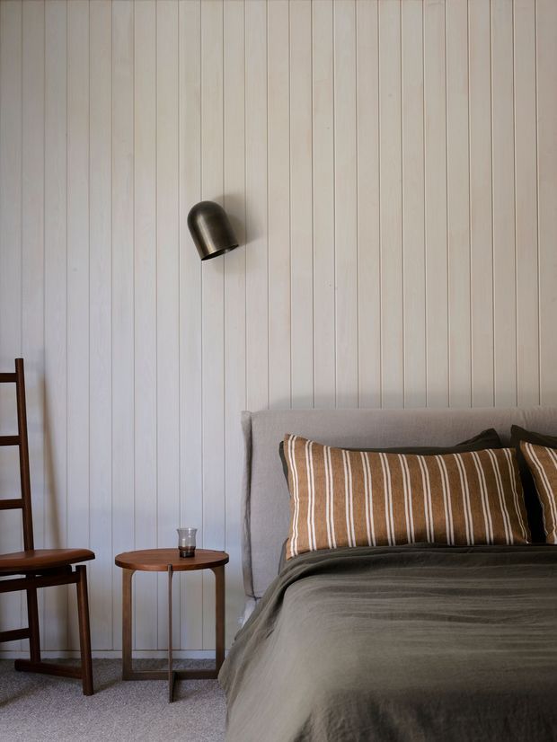 A calm, softly lit bedroom vignette with a bed positioned against a vertically panelled pale wall. The bed has a light upholstered headboard, muted dark grey bedding, and brown-and-cream striped pillows. A dark timber chair and small round timber side table sit to the left, with a compact dark metal wall light above the bed. The restrained natural palette and simple furnishings create a warm, refined atmosphere.