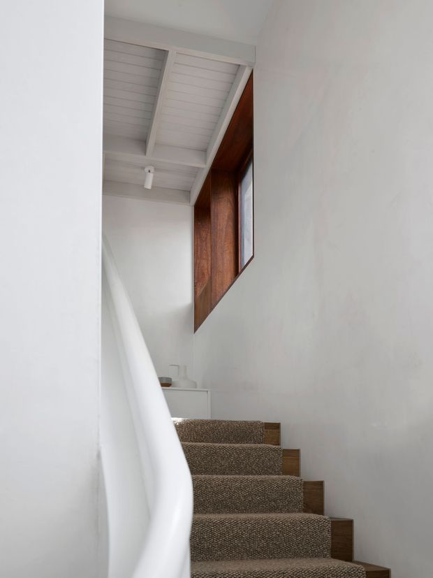A minimalist interior stairway viewed from below, with white painted walls, a white slatted ceiling with exposed beams, a sculptural white balustrade, and brown textured carpeted stairs. A dark timber-clad feature wall and small landing basin are visible above the staircase.