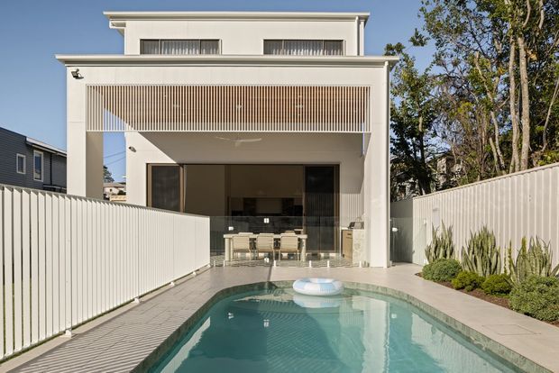 A contemporary two-storey residential facade viewed from a narrow backyard pool area. The white rendered building has strong horizontal roofline trim, a deep covered outdoor entertaining area, large dark-framed glazing, and a vertical timber-look privacy screen above the opening. A long rectangular swimming pool, pale paving, white privacy fences, and landscaped planting occupy the foreground.