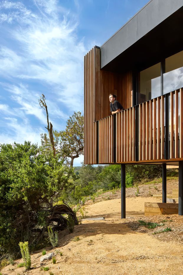 Exterior architectural photograph of a contemporary elevated house set on a dry, vegetated sloping site. The building has a projecting upper level supported on slender dark posts, extensive timber vertical screening and cladding, a dark recessed balcony, large glazing, and a flat concrete roof or parapet. The surrounding landscape is rugged and natural under a bright blue sky.