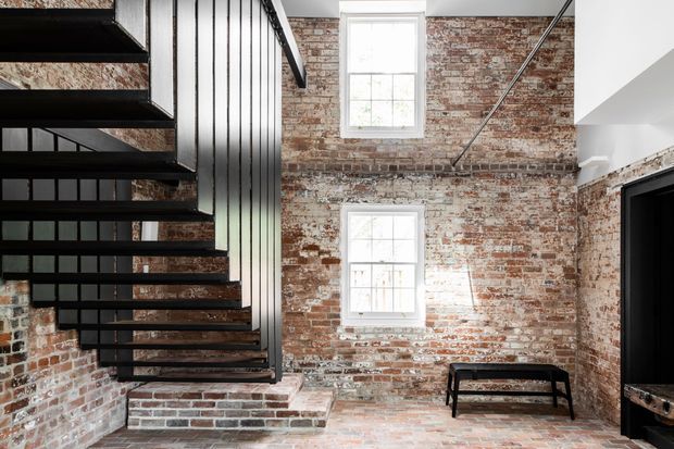 A bright industrial-style interior stairwell with an open-riser black metal staircase, exposed weathered red brick walls, and two white-framed windows. A raised brick platform and low black bench occupy the lower area, while a dark doorway or built-in opening is visible on the right. The space has strong raw-material character, high ceilings, and abundant natural light.