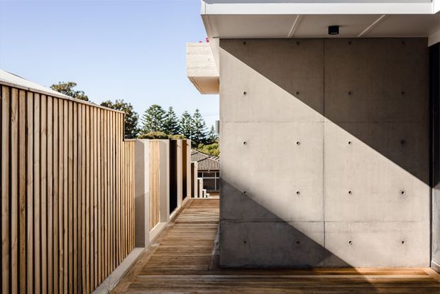 Exterior side passage of a contemporary building, featuring a timber-decked walkway, tall vertical timber privacy fencing, and a large exposed concrete wall with strong geometric sunlight and shadow. The view includes neighbouring rooflines and mature trees in the background.