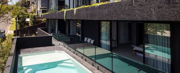 Modern multi-level residential exterior with dark textured rendered walls, extensive floor-to-ceiling glazing, glass balustrades, and a pale turquoise swimming pool terrace. The image emphasizes the building facade and pool area, with balconies and shaded outdoor living spaces visible along the side.
