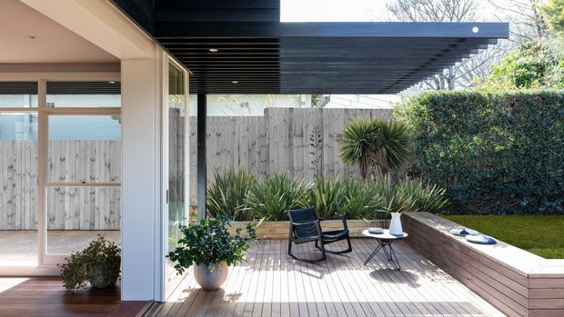 A bright contemporary outdoor deck and patio viewed from beside the house, with pale timber decking, a black slatted pergola, glass-lined indoor-outdoor connection, lounge chairs, a small round table, raised garden beds, hedges and sculptural planting. The space feels open, relaxed and coastal, with glimpses of blue water or sky beyond the glazing.
