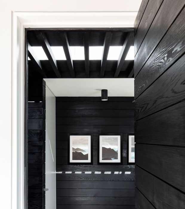 View through an open doorway into a dark, dramatic interior clad in horizontal black-stained timber boards. Exposed dark timber ceiling beams admit narrow strips of daylight, while framed mountain artwork lines the rear wall. White door trim and a simple black cylindrical ceiling light provide contrast.
