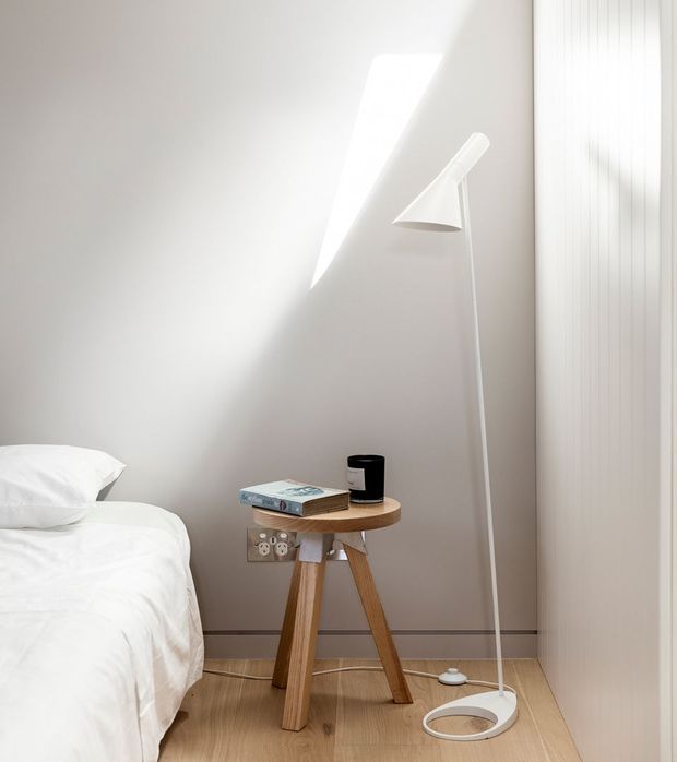 Bright, minimalist bedroom corner with white bedding, pale neutral walls, light timber flooring, a small round timber bedside table, and a slender white floor lamp. Vertical fluted wall panelling or cabinetry appears on the right, while sunlight creates a strong geometric patch of light on the wall.
