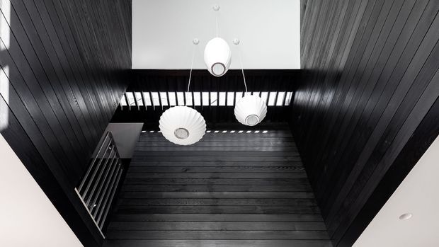 Upward-looking view of a dramatic dark timber-lined double-height interior void, featuring three suspended white pendant lights, clerestory-style glazing that casts striped light, and a black metal stair or balustrade at the left. The space has a high-contrast, minimalist architectural character.