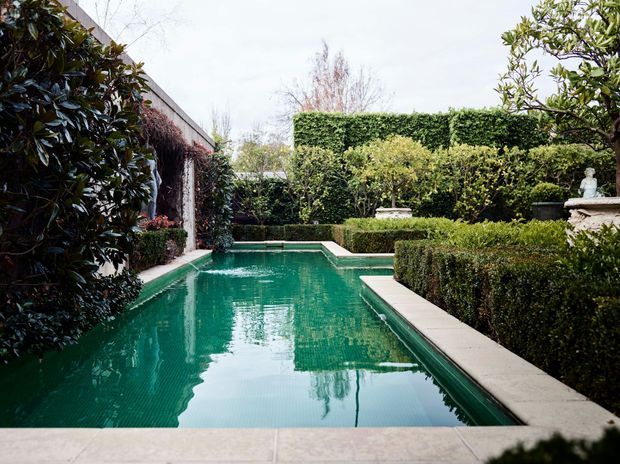 A formal landscaped courtyard centered on a long rectangular swimming pool. The pool is bordered by pale stone paving and clipped box hedges, with dense evergreen planting, tall trimmed hedges, a rendered garden wall, and classical stone ornaments creating a secluded, symmetrical garden setting.