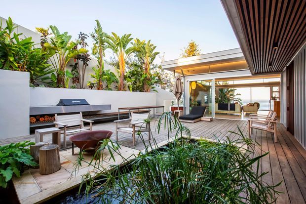 A lush contemporary courtyard entertaining area with timber decking, built-in outdoor kitchen and barbecue, outdoor dining and lounge furniture, and abundant tropical planting. A covered timber-slat ceiling extends along the right side, while the space opens into a bright glazed living area beyond.