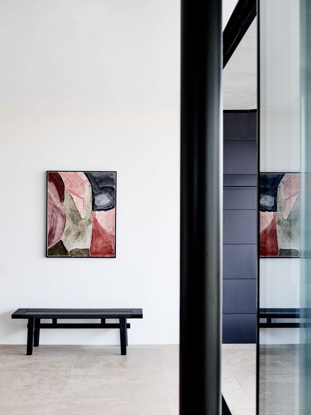 Minimalist interior view of a bright entry or hallway with a white wall, large pale stone or porcelain tiled floor, a simple dark timber bench, and an abstract artwork. A black-framed glazed partition or doorway creates a strong vertical division and reflects the artwork and bench. The space has clean contemporary detailing and a restrained black, white, and pale neutral palette.