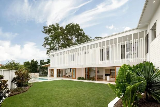 A bright two-storey white residential house viewed from a landscaped backyard. The home has a long upper-level balcony or gallery enclosed by closely spaced vertical white balustrades, white horizontal cladding, large glazed openings, and a low-pitched roof. The ground level opens onto a broad green lawn with a swimming pool at the side and tropical planting around the garden.