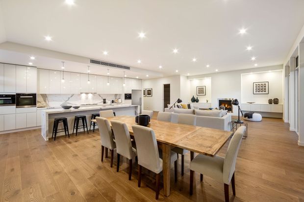A bright, spacious open-plan interior combining a contemporary kitchen, dining area, and living room. The kitchen has sleek white full-height cabinetry, a pale stone island with seating for four, integrated appliances, and a marble-look splashback. A large timber dining table with upholstered chairs occupies the foreground, while the living area includes a sectional sofa, fireplace, and built-in media joinery. Recessed ceiling lighting and warm timber flooring create a refined, inviting atmosphere.