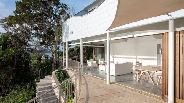 A modern multi-level home with a spacious covered outdoor terrace overlooking a leafy, sloping landscape. The facade features extensive floor-to-ceiling glazing, white horizontal cladding, slender white columns, a curved roofline, and timber screening. The terrace has tiled paving, an outdoor dining setting, built-in or large white seating, and a retractable-style fabric shade.