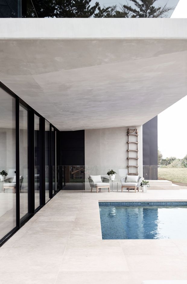 A refined modern poolside terrace beneath a broad projecting concrete roof slab. Floor-to-ceiling black-framed glazing lines the left side, opening onto a minimalist outdoor sitting area with pale upholstered chairs, a small table, and potted plants. Large pale stone or porcelain paving surrounds a rectangular blue-tiled swimming pool, while white and charcoal architectural surfaces create a restrained, high-contrast composition.