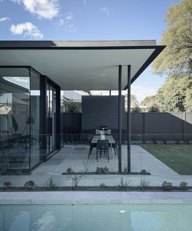 A contemporary residential outdoor entertaining area beside a swimming pool. The covered patio has a broad flat roof with slim dark posts, full-height black-framed glazing, a dining table and chairs, an outdoor barbecue against a dark brick feature wall, and pale stone paving. Linear garden beds, a dark vertical-slat fence, and mature trees surround the space.