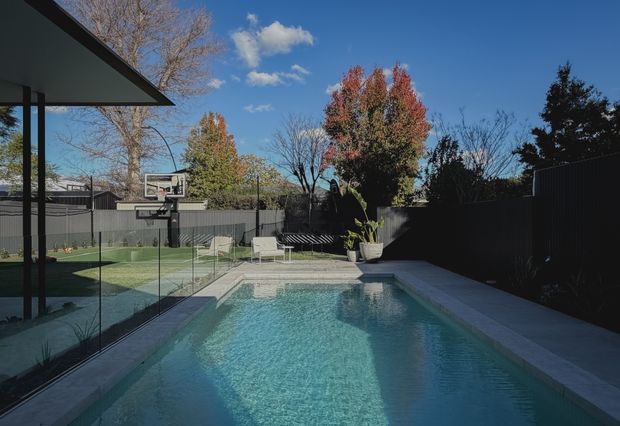 A compact outdoor swimming pool occupies a landscaped backyard courtyard, with pale paving, clear glass pool fencing, dark boundary fencing, potted plants, and a basketball hoop. A covered outdoor structure is visible at left, while mature trees and blue sky form the background. The image has a contemporary residential outdoor setting with strong sunlight and cool blue-green water.