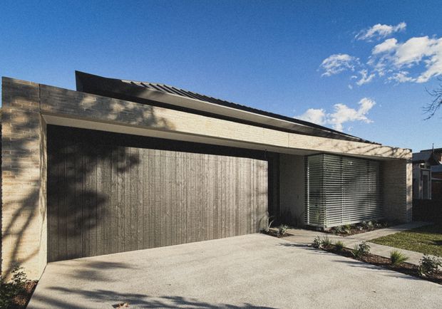 Street-facing view of a compact contemporary single-storey house facade, dominated by a wide timber-clad garage door and matching masonry or stone-toned facade walls. A low-pitched dark roof projects over the garage and entry area, while horizontal louvres screen the front glazing. A concrete driveway and narrow landscaped garden strip complete the foreground.