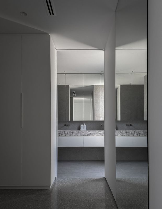A monochromatic contemporary bathroom viewed through a narrow passage, featuring a floating grey vanity with integrated basin, large mirrored wall cabinetry, a marble-patterned splashback, dark grey wall and floor finishes, and tall minimalist grey cabinetry in the foreground. Recessed ceiling lighting and concealed joinery create a clean, highly streamlined interior.